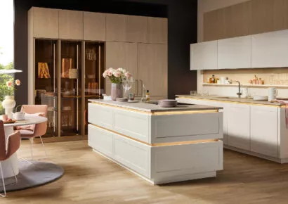 Elegante Nolte Designküche mit klaren Linien, hochwertiger Lackfront in Magnolia und stilvollen Holzakzenten. Die großzügige Kochinsel mit integrierter Beleuchtung trifft auf eine edle Vitrinenlösung und schafft ein modernes, wohnliches Ambiente mit Designanspruch.